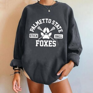 Pu&ograve; includere: Felpa grigio scuro con la scritta bianca "PALMETTO STATE" sopra "STICK BALL" e "FOXES". Il design include bastoni da lacrosse incrociati e un'impronta di zampa. La felpa ha maniche lunghe e girocollo.