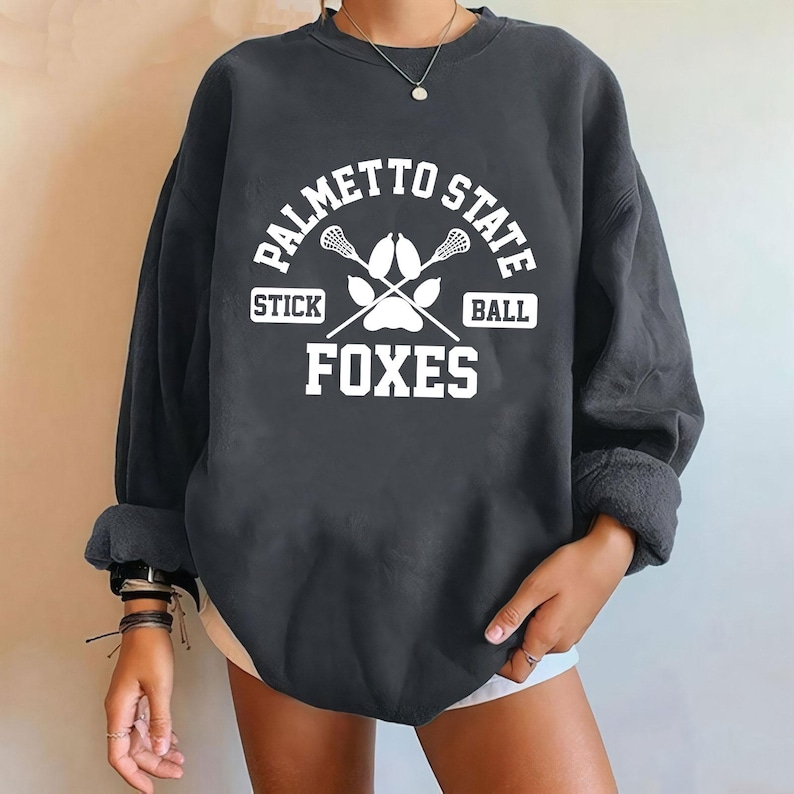 Pu&ograve; includere: Felpa grigio scuro con la scritta bianca "PALMETTO STATE" sopra "STICK BALL" e "FOXES". Il design include bastoni da lacrosse incrociati e un'impronta di zampa. La felpa ha maniche lunghe e girocollo.