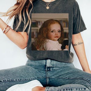 May include: Dark gray t-shirt featuring a framed image of a doll with red hair. The doll is wearing a light pink dress. The person in the image is wearing blue jeans and white sneakers. A gold necklace and bracelet are also visible.