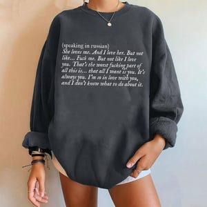 May include: Dark gray sweatshirt with white text. The text reads, "(speaking in russian) She loves me. And I love her. But not like... Fuck me. But not like I love you..." The long-sleeved top has rolled-up sleeves.