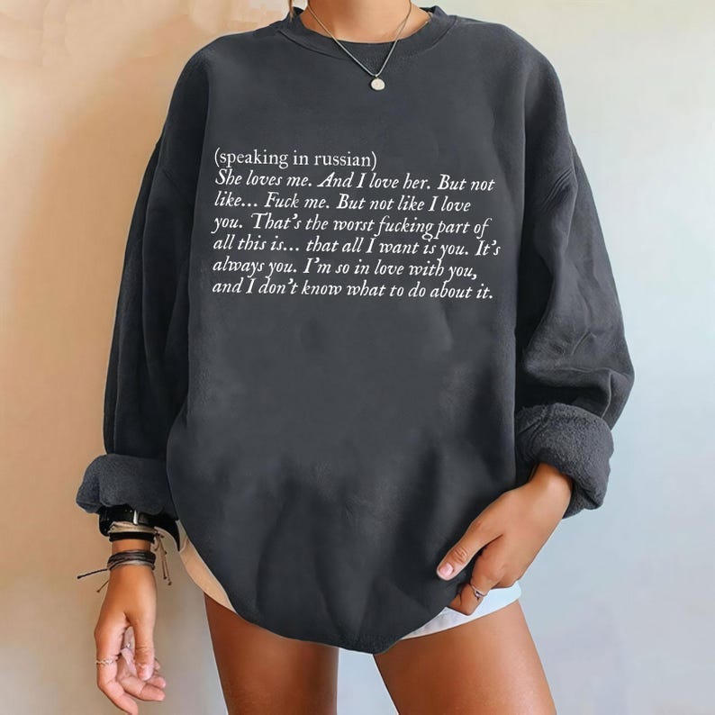 May include: Dark gray sweatshirt with white text. The text reads, "(speaking in russian) She loves me. And I love her. But not like... Fuck me. But not like I love you..." The long-sleeved top has rolled-up sleeves.