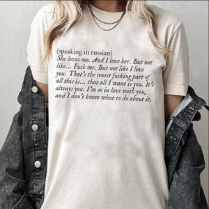 May include: Cream-colored t-shirt with black text. The text includes the phrase "(speaking in russian)" followed by a series of romantic and slightly vulgar statements. The shirt is worn with a denim jacket.