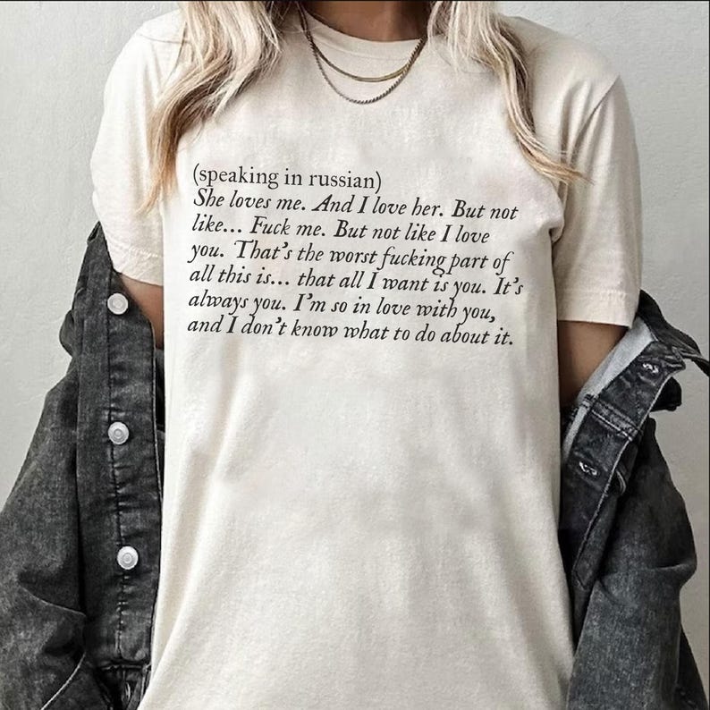 May include: Cream-colored t-shirt with black text. The text includes the phrase "(speaking in russian)" followed by a series of romantic and slightly vulgar statements. The shirt is worn with a denim jacket.