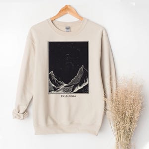 May include: A beige sweatshirt featuring a black and white mountain landscape design within a rectangular frame. The text "Ex Altiora" is printed below the image. The sweatshirt is hanging on a wooden hanger.