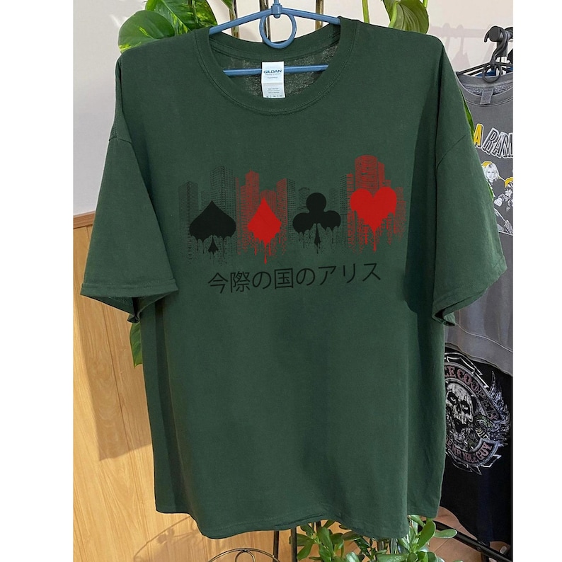 Puede incluir: Camiseta verde oscuro con s&iacute;mbolos de naipes en rojo y negro, y texto japon&eacute;s. El dise&ntilde;o incluye una silueta de paisaje urbano sobre los s&iacute;mbolos. La camiseta est&aacute; colgada en una percha.
