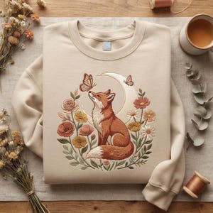 Könnte beinhalten: Ein beigefarbenes Sweatshirt mit Rundhalsausschnitt und einer Stickerei, die einen Fuchs in der Mitte eines Halbmondes zeigt. Der Fuchs ist von einer kreisförmigen Anordnung aus bunten Blumen, Blättern und zwei Schmetterlingen in Orange- und Gelbtönen umgeben.