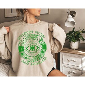 Puede incluir: Sudadera beige con un dise&ntilde;o gr&aacute;fico circular verde que presenta un ojo y las palabras "STATEMENT BEGINS" y "STATEMENT ENDS". El dise&ntilde;o gr&aacute;fico est&aacute; rodeado de varios s&iacute;mbolos. La sudadera es de color neutro.