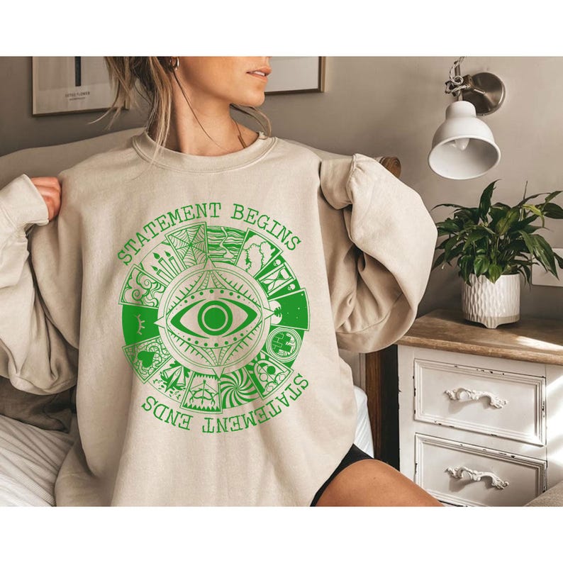 Puede incluir: Sudadera beige con un dise&ntilde;o gr&aacute;fico circular verde que presenta un ojo y las palabras "STATEMENT BEGINS" y "STATEMENT ENDS". El dise&ntilde;o gr&aacute;fico est&aacute; rodeado de varios s&iacute;mbolos. La sudadera es de color neutro.