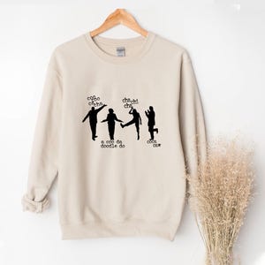 Puede incluir: Sudadera beige con un gráfico negro de cuatro figuras bailando y las frases "coo coo cacha", "cha chi cha", "a coo da doodle do" y "coca caw". La sudadera está colgada en una percha de madera.