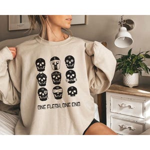 May include: Beige sweatshirt featuring a grid of nine black skull designs, each with unique embellishments. The text "ONE FLESH, ONE END" is printed below the skulls. The sweatshirt is a casual, comfortable style.