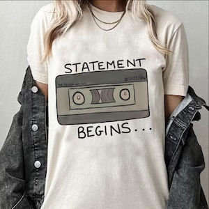Könnte beinhalten: Cremefarbenes T-Shirt mit einer Kassettenband-Grafik und dem Text "STATEMENT BEGINS...". Die Grafik ist in Grau- und Brauntönen gehalten, der Text ist schwarz. Das Shirt trägt außerdem den Text "THE MAGNIFIC INSTITUTE" und die Nummer "#0122204".