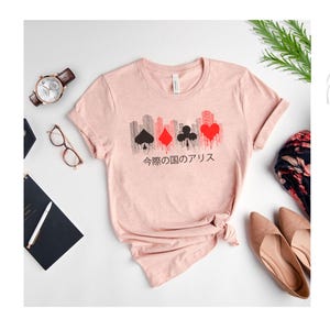 Puede incluir: Una camiseta rosa claro con un dise&ntilde;o de paisaje urbano con s&iacute;mbolos de naipes en negro y rojo. Texto japon&eacute;s impreso debajo. La camiseta se presenta con un reloj, gafas y zapatos.