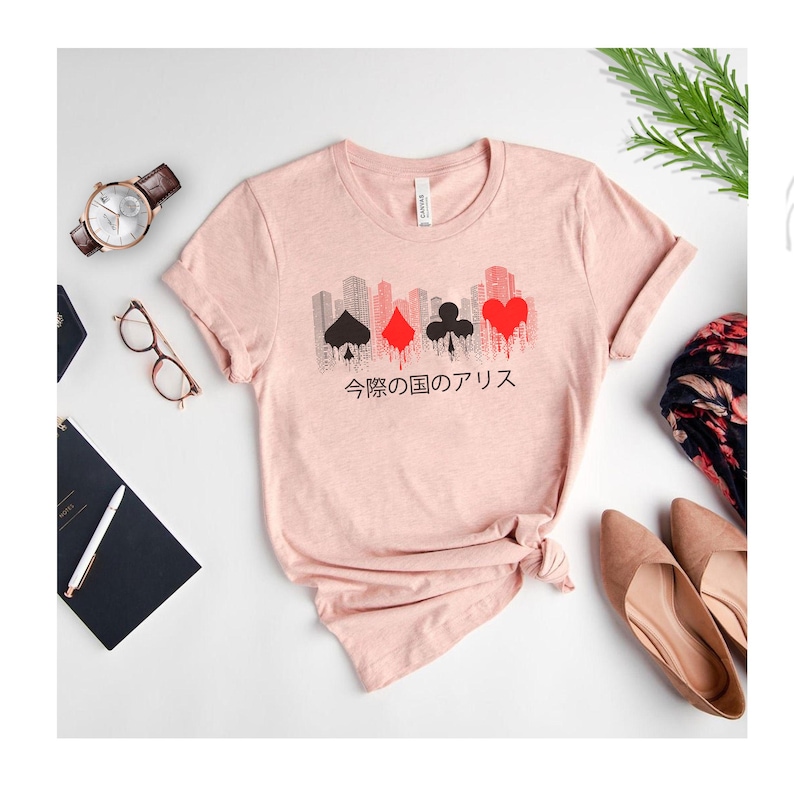 Puede incluir: Una camiseta rosa claro con un dise&ntilde;o de paisaje urbano con s&iacute;mbolos de naipes en negro y rojo. Texto japon&eacute;s impreso debajo. La camiseta se presenta con un reloj, gafas y zapatos.