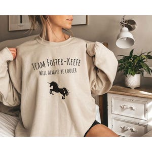 Può includere: Felpa beige con la scritta "Team Foster-Keefe Will Always Be Cooler" e una silhouette di unicorno nero. La felpa ha girocollo e maniche lunghe. L'immagine è di una persona che indossa la felpa.