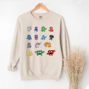 May include: Beige sweatshirt featuring a colorful cartoon graphic of various animated characters. The characters are in different colors and shapes, with expressive faces. The sweatshirt is hanging on a wooden hanger.
