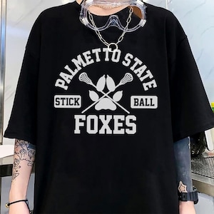 Pu&ograve; includere: T-shirt nera con grafica bianca. La grafica include il testo "PALMETTO STATE", bastoni da lacrosse incrociati, un'impronta di zampa e le parole "STICK BALL" e "FOXES". La maglietta ha maniche corte.