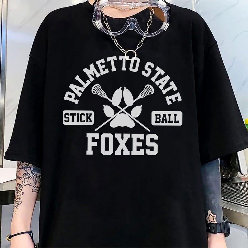 Pu&ograve; includere: T-shirt nera con grafica bianca. La grafica include il testo "PALMETTO STATE", bastoni da lacrosse incrociati, un'impronta di zampa e le parole "STICK BALL" e "FOXES". La maglietta ha maniche corte.
