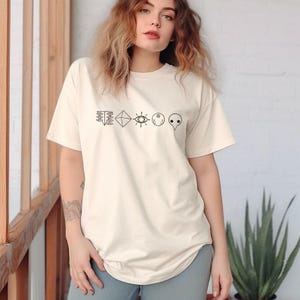 Könnte beinhalten: Cremefarbenes T-Shirt mit einer Reihe schwarzer Strichzeichnungen. Die Symbole umfassen geometrische Formen, ein Auge, einen Mond und ein stilisiertes Gesicht. Das Shirt hat eine lässige, übergroße Passform.