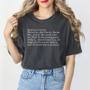May include: Dark gray t-shirt with white text. The text reads: "(speaking in russian) She loves me. And I love her. But not like... Fuck me. But not like I love you. That's the worst fucking part of all this is... that all I want is you. It's always you. I'm so in love with you, and I don't know what to do about it."