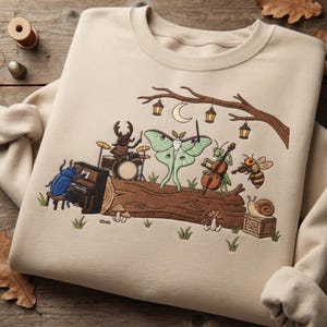 Könnte beinhalten: Ein beigefarbenes Sweatshirt mit Rundhalsausschnitt und einer Stickerei von Insekten, die auf einem Baumstamm Musikinstrumente spielen. Ein Käfer spielt Klavier, ein Hirschkäfer Schlagzeug, ein Falter dirigiert, eine Gottesanbeterin Cello, eine Biene Geige und eine Schnecke sitzt auf einer Kiste.