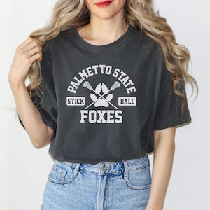 Pu&ograve; includere: T-shirt grigio scuro con la scritta "PALMETTO STATE" in alto e "STICK BALL" su entrambi i lati di un grafico di bastone da lacrosse e impronta di zampa. La parola "FOXES" &egrave; stampata sotto.