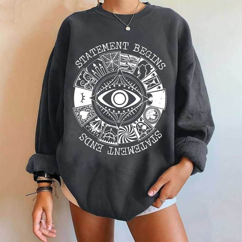 Puede incluir: Sudadera gris oscuro con un dise&ntilde;o gr&aacute;fico blanco. El dise&ntilde;o presenta un ojo grande en el centro, rodeado por un patr&oacute;n circular con las palabras "STATEMENT BEGINS" y "STATEMENT ENDS". Las mangas est&aacute;n dobladas.