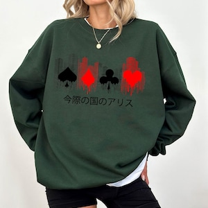 Puede incluir: Sudadera verde oscuro con un gr&aacute;fico de palos de cartas en negro y rojo, y una silueta de paisaje urbano. Texto japon&eacute;s impreso debajo. La sudadera tiene cuello redondo y mangas largas.