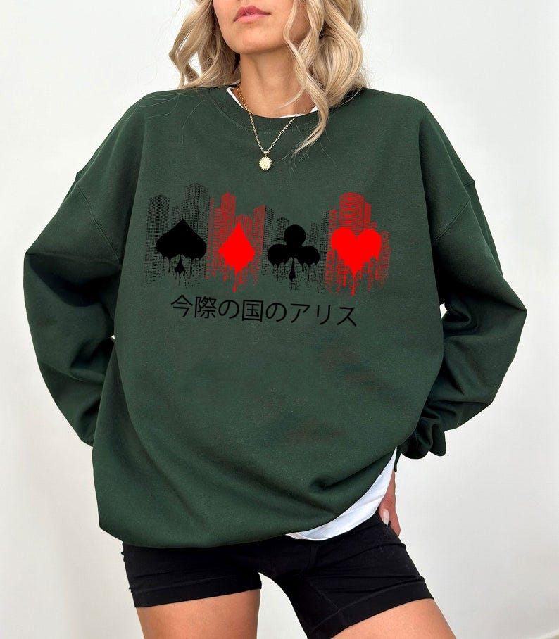Puede incluir: Sudadera verde oscuro con un gr&aacute;fico de palos de cartas en negro y rojo, y una silueta de paisaje urbano. Texto japon&eacute;s impreso debajo. La sudadera tiene cuello redondo y mangas largas.