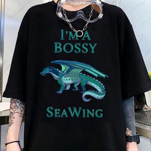 May include: Black t-shirt with the text "I'M A BOSSY SEA WING" in teal. A teal and green dragon graphic is centered below the text. The shirt has short sleeves.