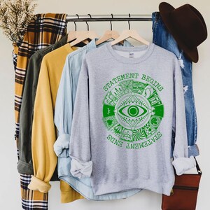 Puede incluir: Una sudadera gris con un dise&ntilde;o gr&aacute;fico verde que presenta un ojo y las palabras "STATEMENT BEGINS" y "STATEMENT ENDS". La sudadera est&aacute; colgada en una percha con otras prendas, incluyendo una camisa amarilla y una camisa vaquera.