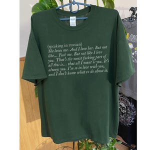 May include: Dark green t-shirt with white text. The text reads "(speaking in russian) She loves me. And I love her. But not like... Fuck me. But not like I love you..." A casual, unisex tee.