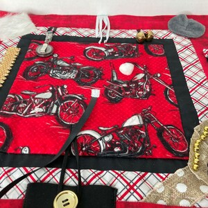May include: A red sensory activity mat with a motorcycle print. The mat features various textures and interactive elements, including zippers, ribbons, and a button. Additional elements include a fuzzy patch, bells, and a small gray felt shape.