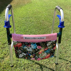 May include: A silver metal walker featuring a black, pink, and blue floral fabric pouch attached to the front crossbar. The pouch has a pink top band with a white label that reads Have a Great Day in black text. The walker has blue plastic accents.