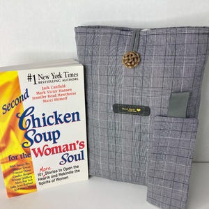 May include: A book titled "A Second Chicken Soup for the Woman's Soul" is next to a gray plaid book sleeve. The book sleeve has a small pocket and a decorative button. The book cover is yellow and white.