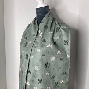 May include: A sage green scarf with a repeating arrow pattern in black, white, and olive green. The scarf is draped over a mannequin, showcasing the fabric's texture and design. This accessory is suitable for adding a touch of style to any outfit.
