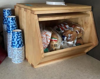 May include: A wooden bread box with a hinged top and a pull-down door. The box is made of light-colored wood and is filled with packaged bread. Blue patterned paper cups are stacked to the left. The text "UrbanDecorNest" is visible.