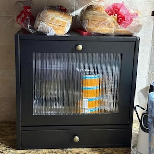 May include: A black bread box with a glass door and a drawer. The door has a ribbed design. Two bags of hamburger buns sit on top of the box. The buns are in clear plastic bags.