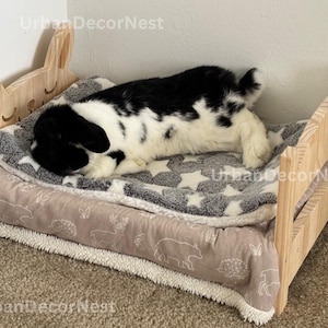 May include: A wooden pet bed with a bunny ear headboard. The bed has a gray and white star-patterned blanket and a light brown and white patterned blanket. A black and white pet is lying on the bed.
