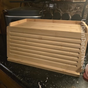May include: A natural bamboo bread box with a slatted design. The box has a flat top and a series of horizontal slats on the front, providing ventilation. The bread box is designed to keep bread fresh and is a kitchen storage solution.