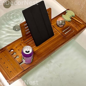 May include: A wooden bathtub tray with a black tablet, a purple can, and a green bath sponge. The tray has cutouts for a drink and other items. The tray is placed over a white bathtub filled with water.