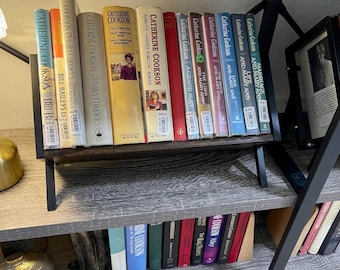 May include: A dark wooden bookshelf with black metal supports, displaying a collection of hardcover books. The books have various colors and titles, including works by Catherine Cookson. A framed picture is also on the shelf.