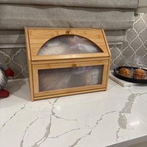 May include: A bamboo bread box with a mesh front and top. The box has a curved top and a small wooden knob. The bread box is sitting on a white countertop.