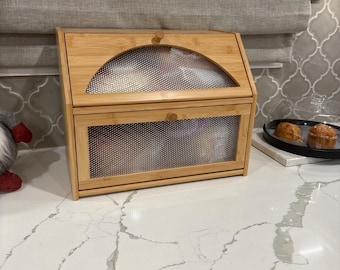 May include: A bamboo bread box with a mesh front and top. The box has a curved top and a small wooden knob. The bread box is sitting on a white countertop.