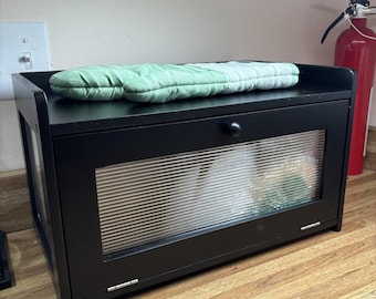 May include: A black wooden bread box with a glass front and a small round knob. A green and white patterned oven mitt rests on top. The bread box is on a wooden surface.