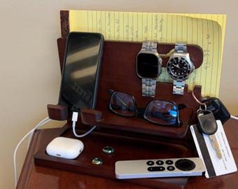 May include: A dark wood bedside organizer with a phone, two watches, glasses, and a remote. A pair of white earbuds, two green glass beads, keys, and a credit card are also present. A yellow notepad is in the back.