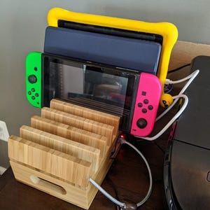 May include: A wooden charging station for a gaming console with a slot for the console and controllers. The console is black with green and pink controllers. A yellow case protects the console. The text "Urban Decor Nest" is visible.