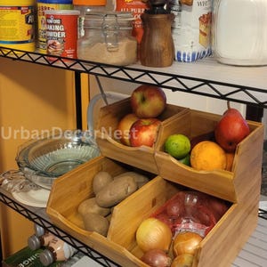 May include: A wooden produce storage bin with three compartments, filled with apples, potatoes, onions, and other fruits and vegetables. The bin sits on a wire shelf, with various pantry items in the background.
