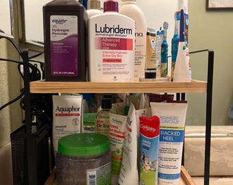 May include: A two-tiered wooden shelf with black metal supports holds various personal care products. Items include bottles of lotion, toothpaste, and a bottle of hydrogen peroxide. The shelf is in a bathroom setting.