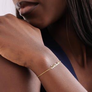 Può includere: Un delicato braccialetto color oro con il nome "محمد" in scrittura araba. Il braccialetto è indossato su un polso, con la catena e la targhetta con il nome ben visibili. Lo sfondo è di un tono morbido e neutro.