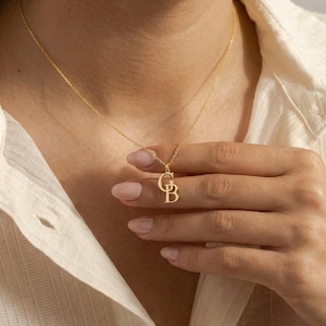 May include: A gold-coloured necklace with a pendant featuring the letters "CB". The pendant hangs from a delicate chain. The necklace is displayed against a neutral background.